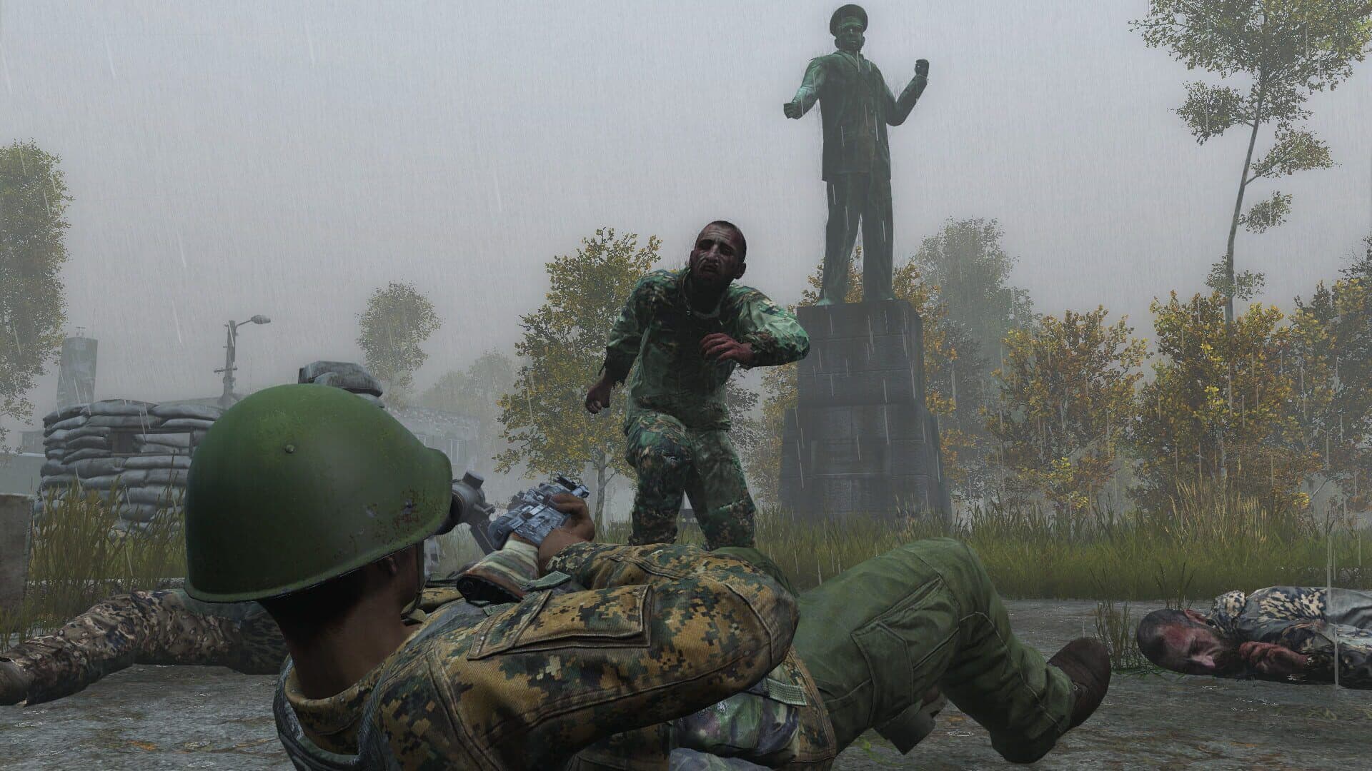 A DayZ survivor lies prone in the rain aiming at an infected in a Chernarus town square, with a body on the ground nearby.